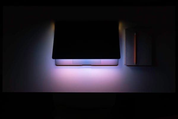 Laptop on a dark desk with soft purple and blue ambient lighting, a notebook and pencil resting beside it.
