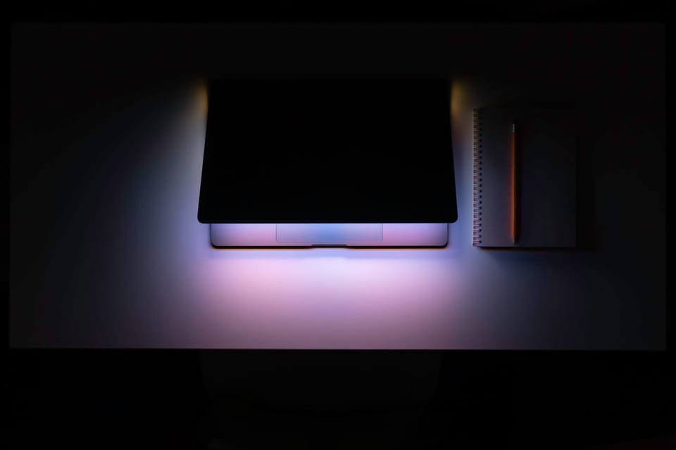 Laptop on a dark desk with soft purple and blue ambient lighting, a notebook and pencil resting beside it.